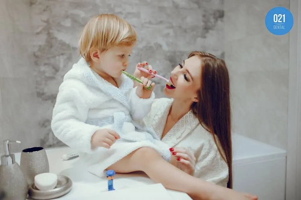 Imagem de uma mãe escovando os dentes com seu filho em um banheiro, ambos usando roupões brancos