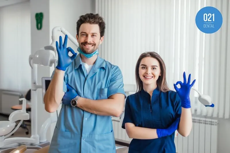 dois dentistas, um homem e uma mulher, com luvas azuis fazendo sinal de ok com a mão dentro de um consultório odontológico