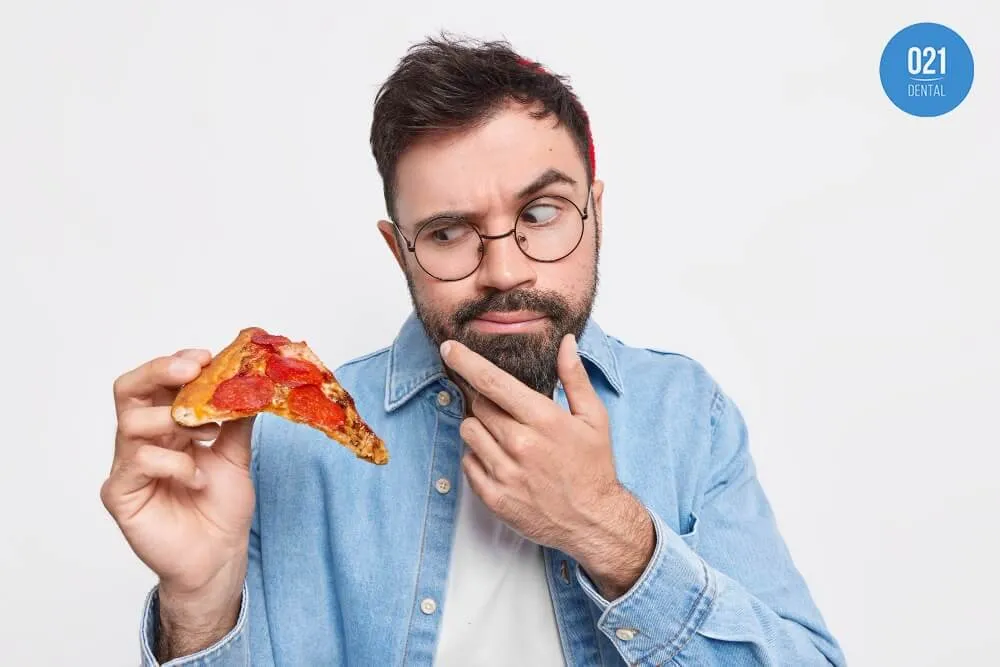 Homem jovem com óculos e jaqueta jeans segurando um pedaço de pizza