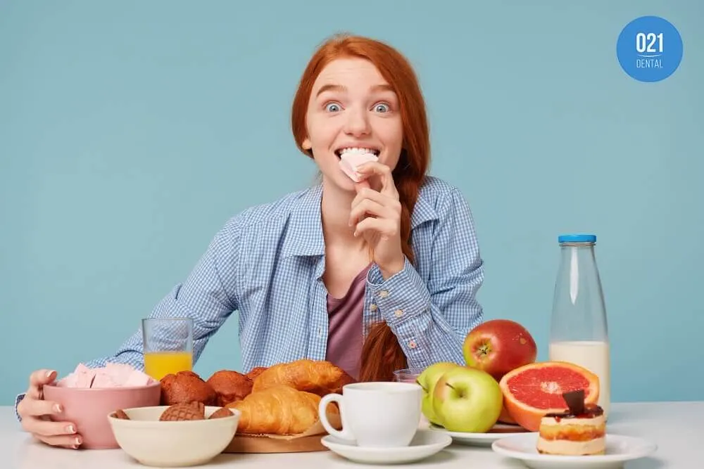 Mulher ruiva com trança lateral e uma camisa azul claro que está mordendo um pedaço de comida enquanto está sentada à mesa com diversos alimentos como frutas, bolos, croissants e suco.
