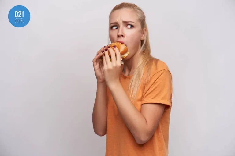 Mulher de blusa laranja comendo hambúrguer olhando para o lado