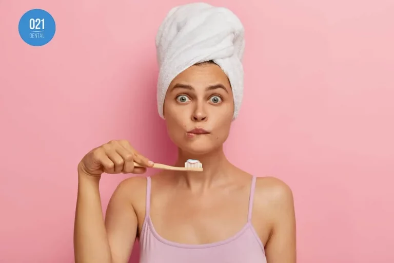 Mulher ao centro da imagem com uma toalha enrolada na cabeça e segurando uma escova de dentes enquanto faz uma careta. Ela está com uma regata rosa e posicionada na frente de um fundo também rosa.