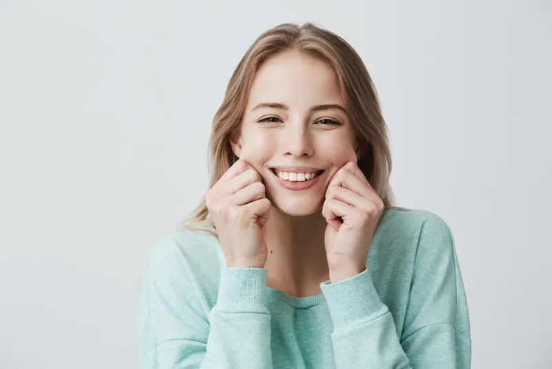 Imagem de uma mulher loira jovem sorrindo segurando as bochechas